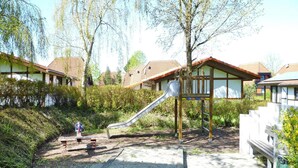Een speeltuin met een glijbaan en een kleine bank, omgeven door groen en woongebouwen.
