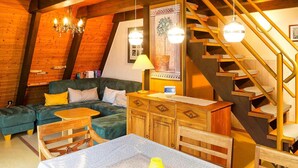 Een knus loft-achtig vertrek met houten plafond, een trap, een bank, een eettafel en een kroonluchter.