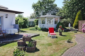 Un jardin agrémenté d’un kiosque, d’une chaise rouge et d’une table blanche recouverte d’une nappe blanche.