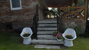 Ein Backsteinhaus mit einer Steintreppe, die zu einer Veranda führt. Auf jeder Seite der Stufen stehen jeweils zwei Korbstühle mit Blumentöpfen.