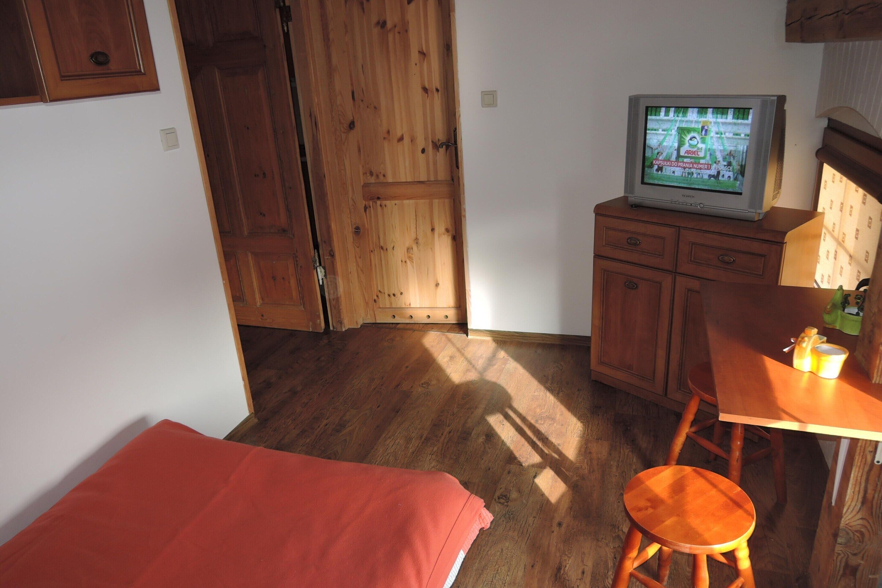 A room with a wooden floor, a bed with a red blanket, a wooden table with a chair, and a television on a wooden cabinet.