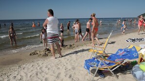 Ein Strand mit badenden und promenierenden Menschen. Liegestühle und Sonnenschirme liegen im Sand.