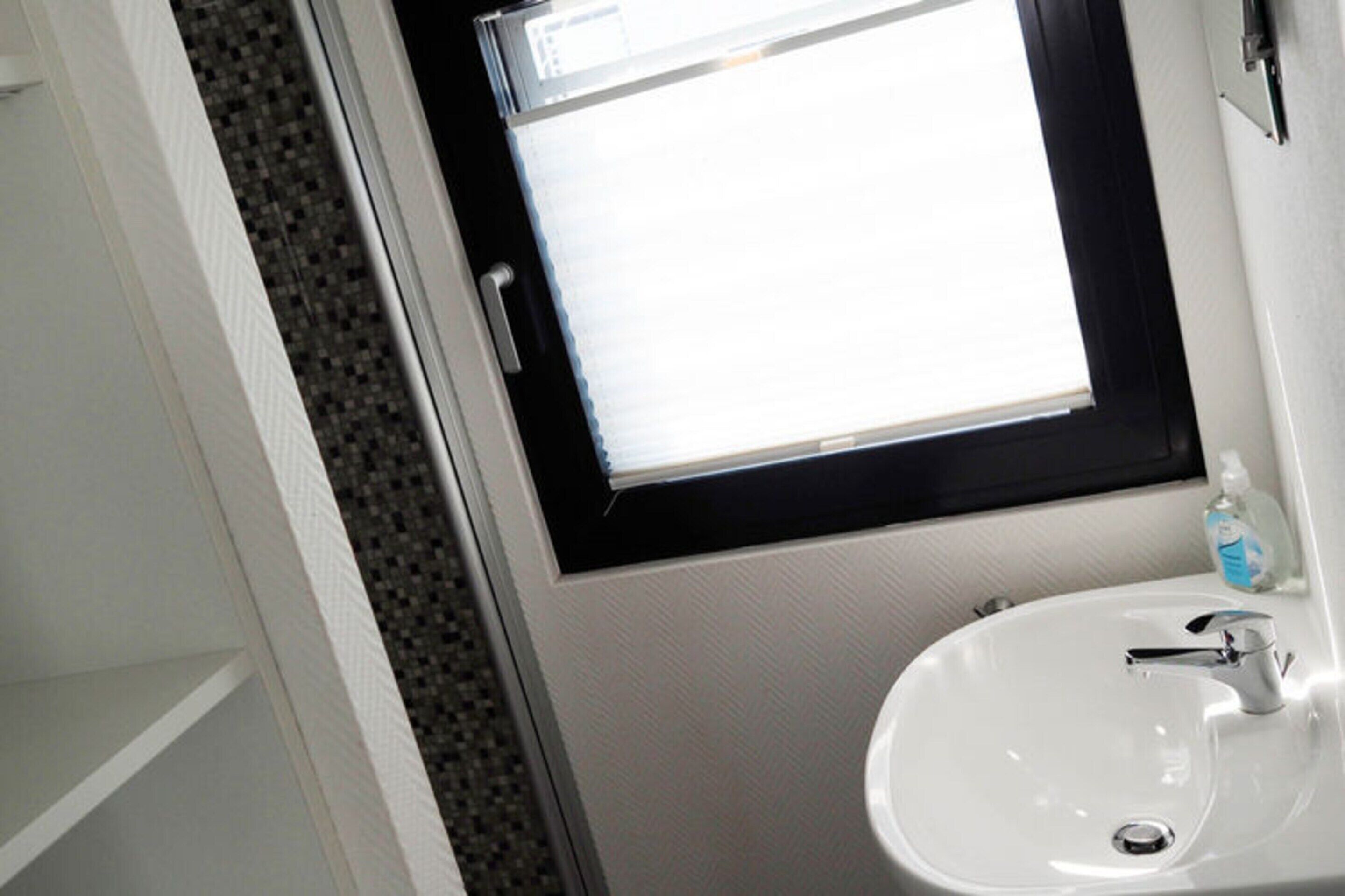 A bathroom with a white sink, a window with a black frame, and a bottle of hand soap on the sink.