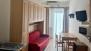 A compact room with a wooden wardrobe, a red sofa, a dining table with chairs, a TV, and a microwave. - Pietra Ligure Beach Residence for 4 (Pietra Ligure)