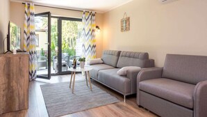 A modern living room with a grey sofa, a wooden side table, and a glass door leading to an outdoor area. - Comfortable Apartments Close to the Beach Darlowko (Darlowo)