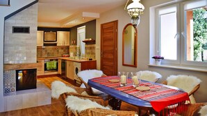Zona pranzo con tavolo apparecchiato per quattro, circondato da sedie in vimini e coperte in pelliccia bianca. Si intravede sullo sfondo una cucina con elettrodomestici moderni e parete di fondo rivestita in piastrelle.
