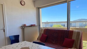 Una habitación con una mesa, un sofá, una silla, un radiador, un reloj y una ventana con vistas a edificios.