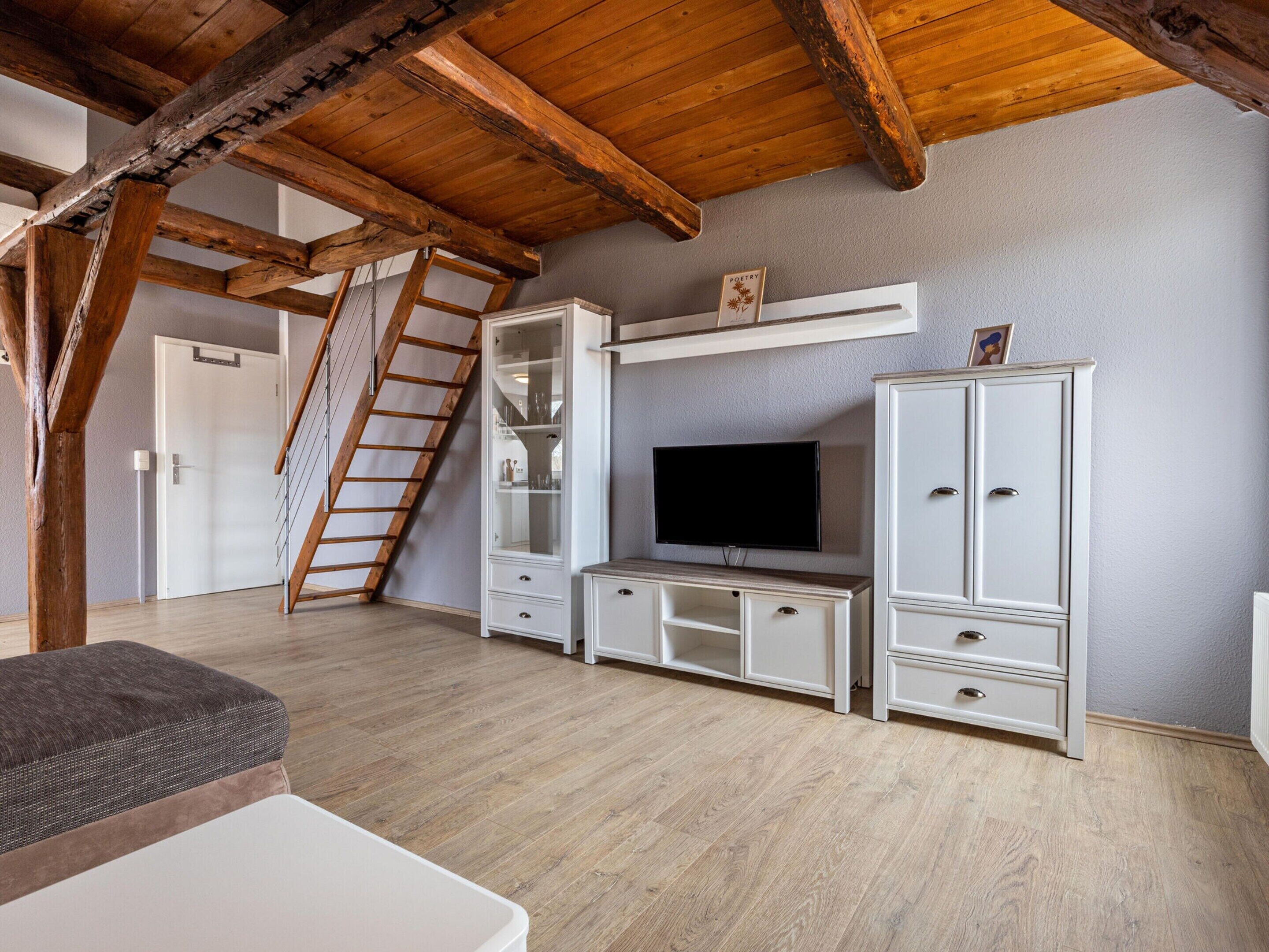 Una stanza in stile loft con travi a vista in legno, una TV fissata a un mobile e una scala che porta a una porta.