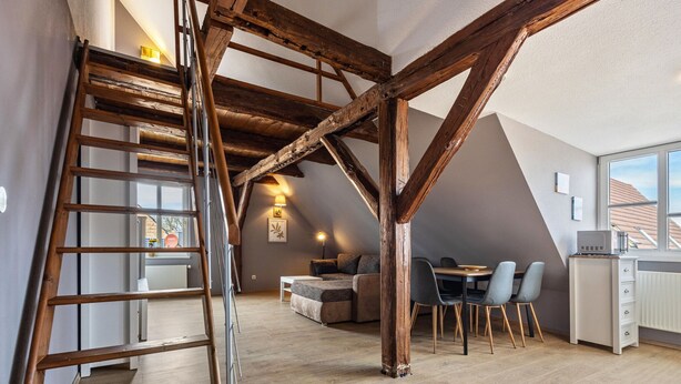 Habitación estilo loft con vigas de madera expuestas, cama en altillo, zona de comedor con sillas y área de cocina con microondas.