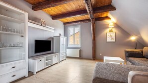 Una sala de estar moderna con techo de madera, televisor, muebles blancos y ventana con vista a un edificio de ladrillo.