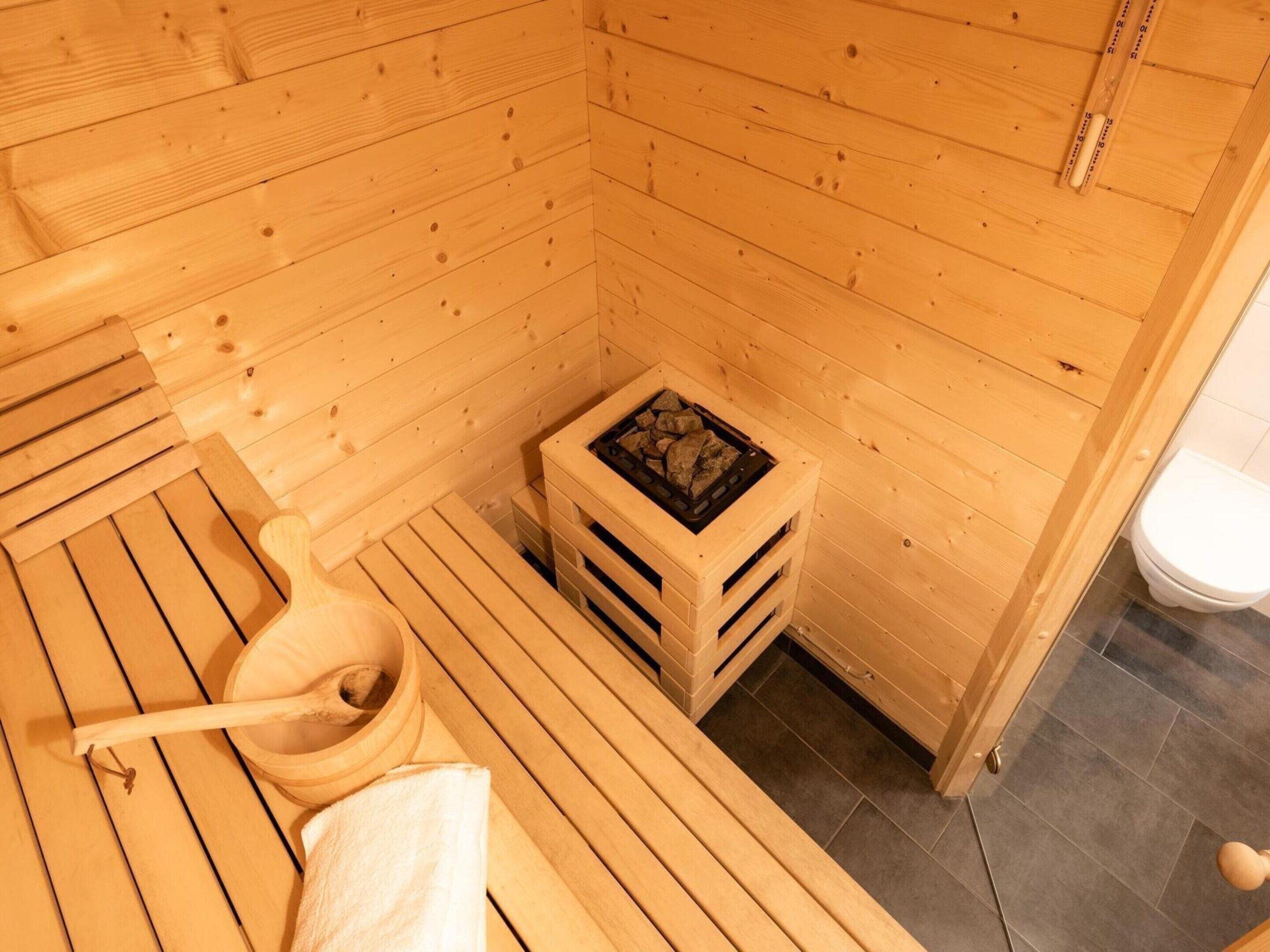 Puuinen sauna, jossa on penkki, puinen ämpäri ja pieni puinen laatikko, jonka sisällä on kiviä.