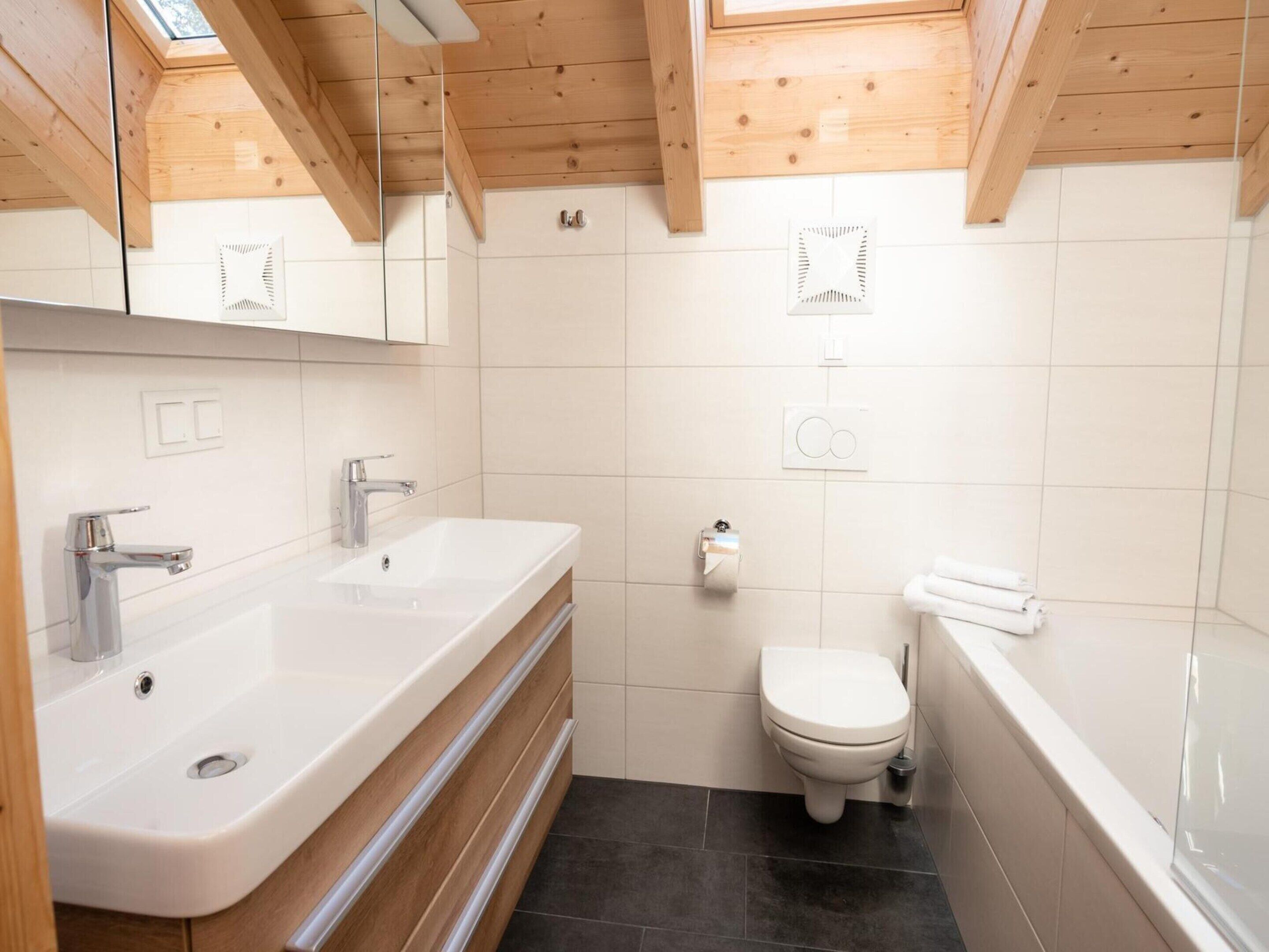 Une salle de bain moderne avec un plafond en bois, des carreaux blancs, un double vasque, des toilettes et une baignoire.