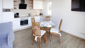 A compact kitchen with white cabinets, a dining table with chairs, and a flat-screen TV mounted on the wall. - Ferienhaus Seeblick, Furstenwerder (Nordwestuckermark)