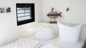 Un lit bien fait, avec du linge de lit blanc, une petite fenêtre à cadre noir et un porte-serviettes mural en forme de poisson.