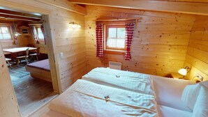 A cozy cabin room with a bed, a small table, and a chair. There is a window with curtains, and a lamp on the nightstand.
