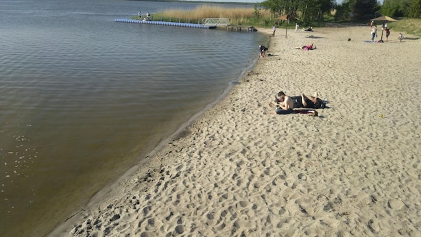 Ein sandiger Strand mit einem Steg, der sich ins Wasser erstreckt, Bäume im Hintergrund und Menschen, die sich in der Sonne entspannen.