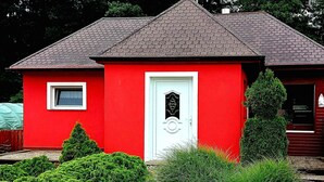 Ein rotes Haus mit einer weißen Tür, einem kleinen Fenster und einem Garten mit Büschen und einer Statue.