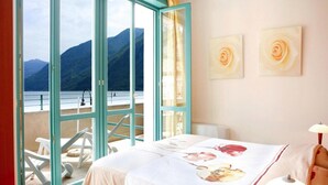 A hotel room with a bed, a red chair, a lamp, and a view of mountains through a glass door. - 4-guest Porlezza Apartment Near Lake Lugano (Porlezza)