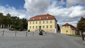Un edificio storico con facciata gialla e tetto rosso, immerso in un selciato e circondato da una recinzione in rete metallica.
