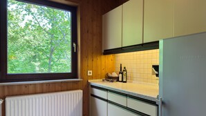 A kitchen with wooden walls, a window with a view of trees, a white radiator, and a stainless steel refrigerator. - Ferienhaus Sackpfeifenblick, Hatzfeld (Hatzfeld)