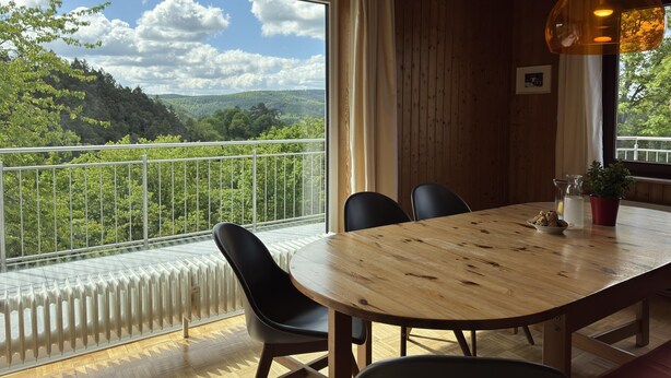 A dining area with a wooden table and chairs, a view of a forest through a large window, and a hanging light fixture. - Ferienhaus Sackpfeifenblick, Hatzfeld (Hatzfeld)