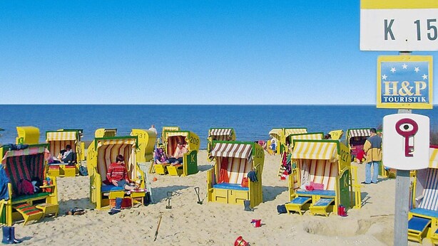 Una spiaggia con casette colorate disposte in sequenza, un cielo azzurro limpido e un cartello per H&P Touristik.