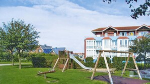 Un'area giochi con altalene e scivolo, circondata da erba e alberi, con un edificio sullo sfondo.
