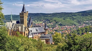 A historic building with multiple towers and a prominent spire, surrounded by lush green hills and a small town below. - Ferienwohnung Remise, Wernigerode (Wernigerode)