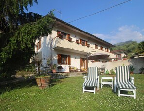 A two-story building with a flat roof, a balcony with potted plants, and a patio area with two striped chairs and a table. - Holiday Home in Camaiore With Mountain Views (Camaiore)