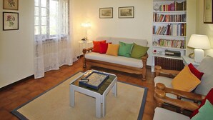 A living room with a sofa, a coffee table, a bookshelf, and a window with lace curtains. - Holiday Home in Camaiore With Mountain Views (Camaiore)