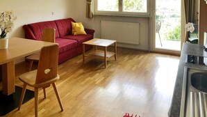 Uma sala de estar acolhedora com um sofá vermelho, mesa de jantar de madeira e cadeiras. Há uma janela com cortinas, um radiador e vista para o verde lá fora.