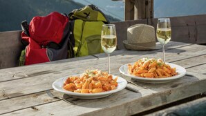 Uma mesa de madeira com dois pratos de massa e dois copos de vinho branco, uma mochila vermelha e uma mochila verde em um banco de madeira com vista para as montanhas ao fundo.