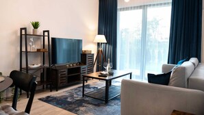 En moderne stue med sofa, fjernsyn og sofabord. Der er et bogreol med dekorative genstande og en lampe.