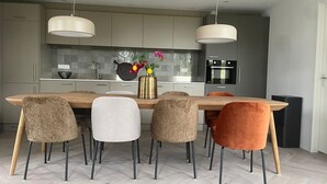 A modern kitchen with a wooden dining table, six chairs, pendant lights, and a vase with flowers.