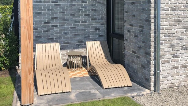 A modern outdoor seating area with two lounge chairs and a small round table, set against a backdrop of a brick wall and a wooden post.