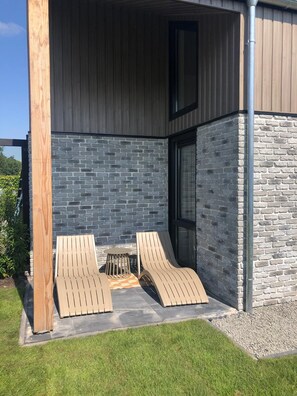 A modern outdoor seating area with two lounge chairs and a small round table, set against a backdrop of a brick wall and a wooden post. - Modern Detached Villa (Tzummarum)