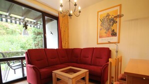 A red sofa, a wooden coffee table, and a chair in a room with a large window, a chandelier, and a framed painting. - Ferienwohnung im Haus Schiefertal, Wieda (Walkenried)