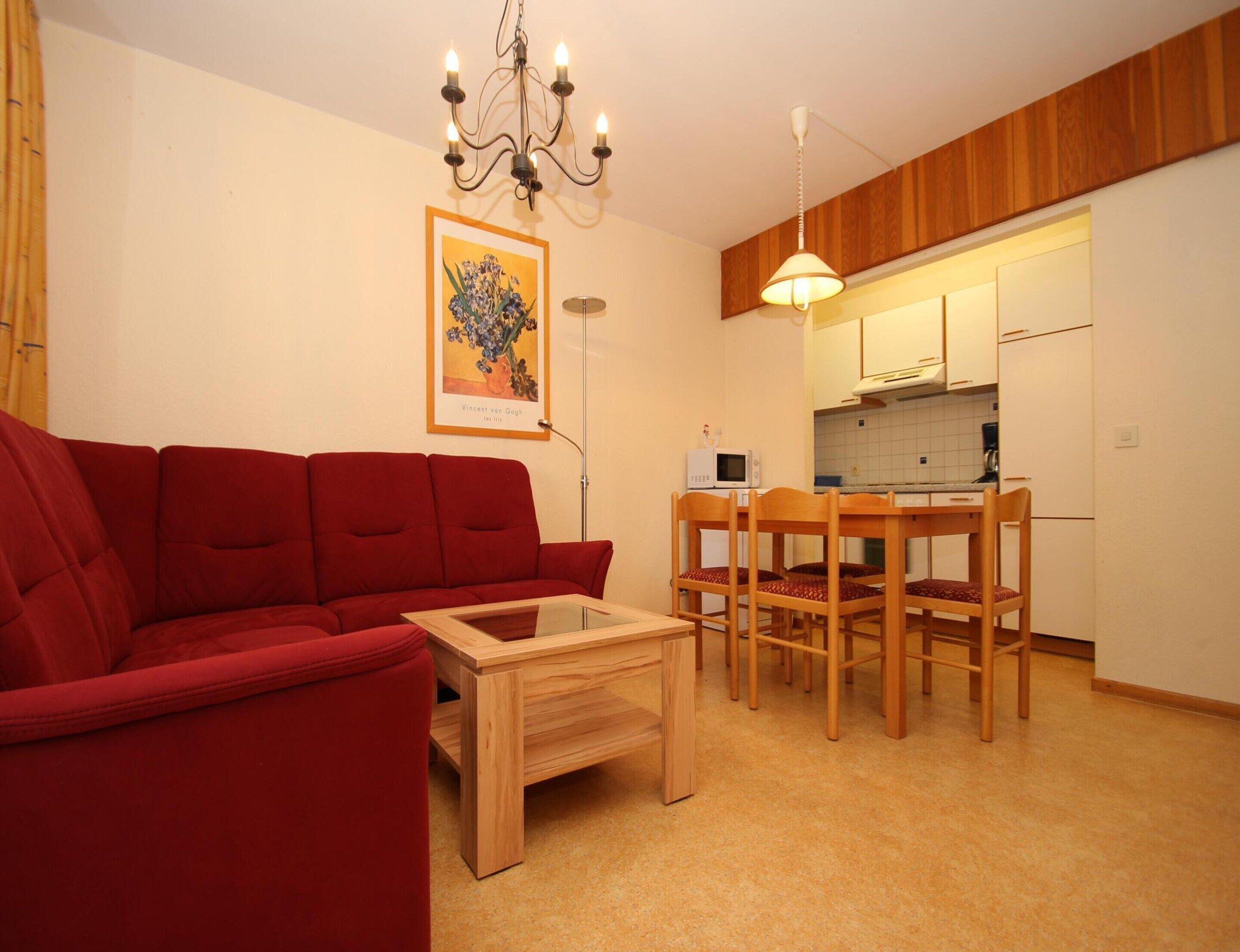 A compact living space with a red sofa, a wooden coffee table, a dining table with chairs, a kitchen area, and a chandelier.