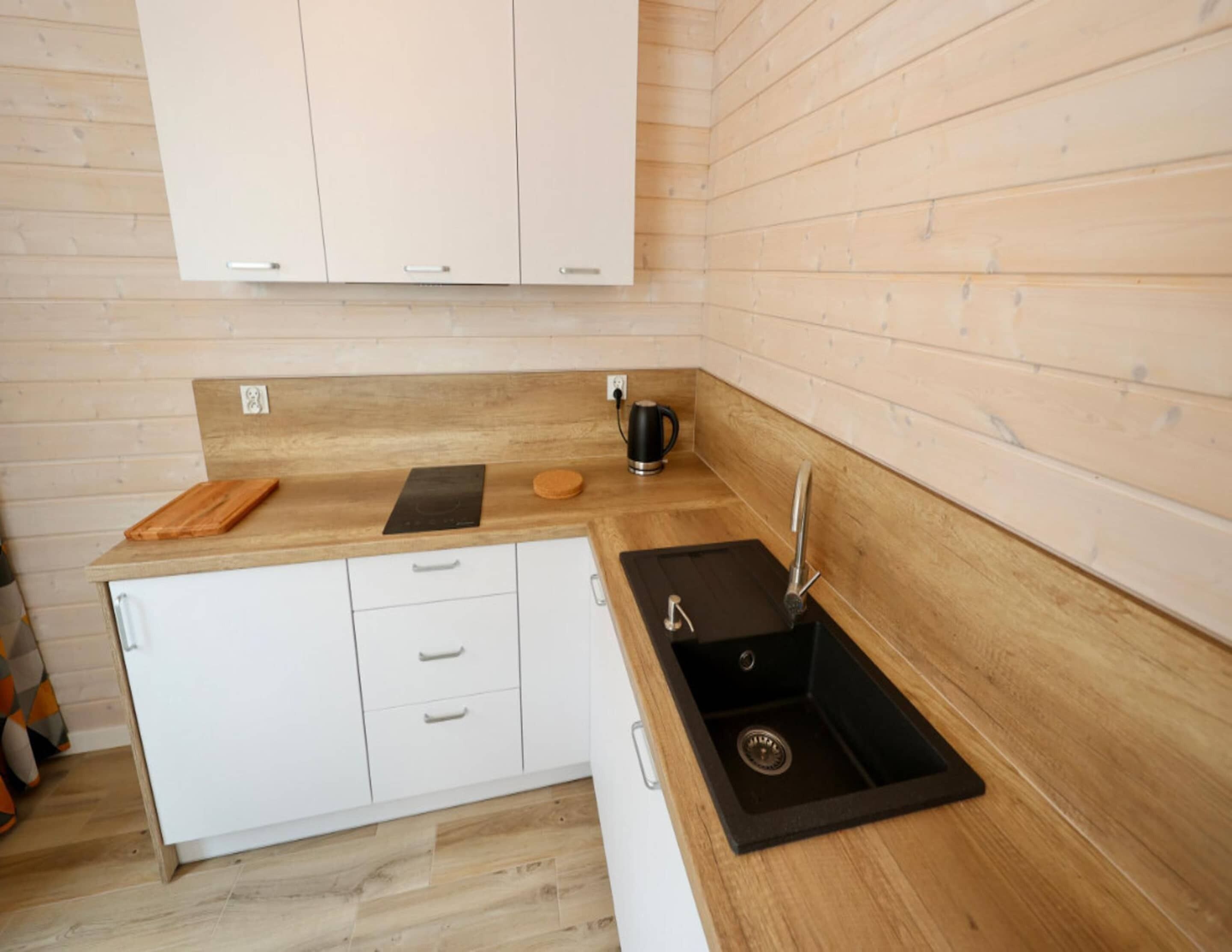 A compact kitchen with wooden cabinetry, a black sink, and a countertop with a cutting board and a kettle.