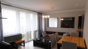 A modern living room with a sofa, dining table, and chairs. There is a large window with curtains, a flat-screen TV mounted on the wall, and a small table with a lamp. - Condo in Swinoujscie Near Baltic Sea Beach (Swinoujscie)