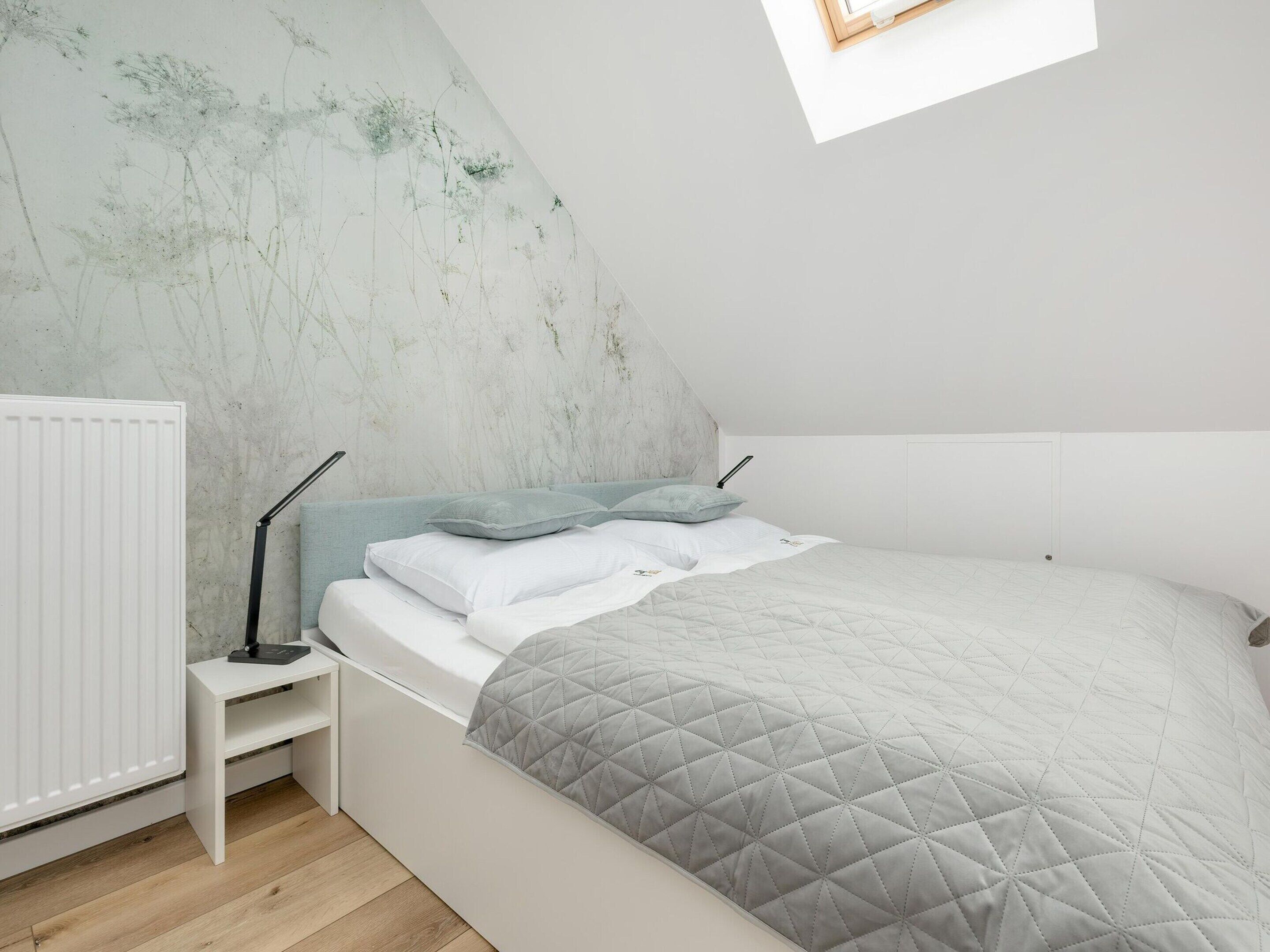 A bedroom with a sloped ceiling, a white bed with a quilted grey blanket, a bedside table with a lamp, and a radiator.