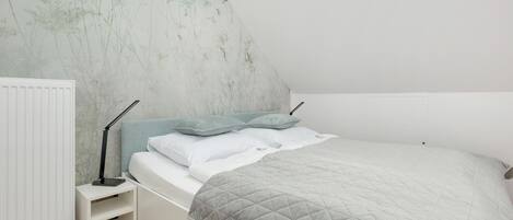 A bedroom with a sloped ceiling, a white bed with a quilted grey blanket, a bedside table with a lamp, and a radiator.