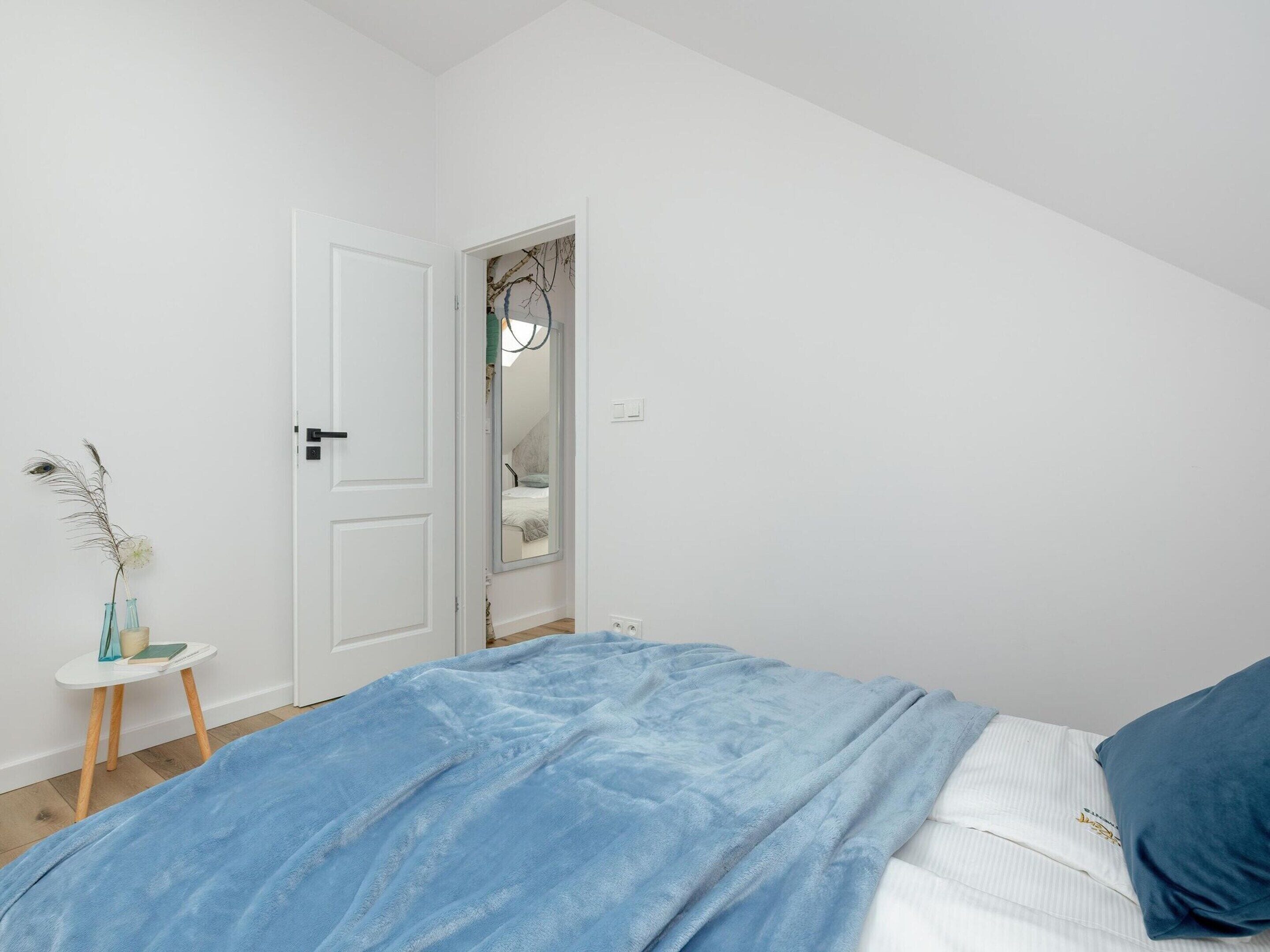 Ein Schlafzimmer mit einem Bett, einer weißen Tür, einem kleinen Tisch mit Vase und Buch sowie einem Spiegel an der Wand.