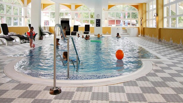 Una piscina interna con pavimento a scacchi, ambiente luminoso e diverse poltrone relax.