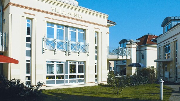Un edificio con el nombre "Villa Sonja" en la fachada, que cuenta con ventanas grandes y un balcón con celosía.
