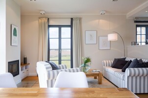 Ein modernes Wohnzimmer mit einer Couch, einem Sessel und einem Couchtisch, einem Kamin und großen Fenstern mit Vorhängen.