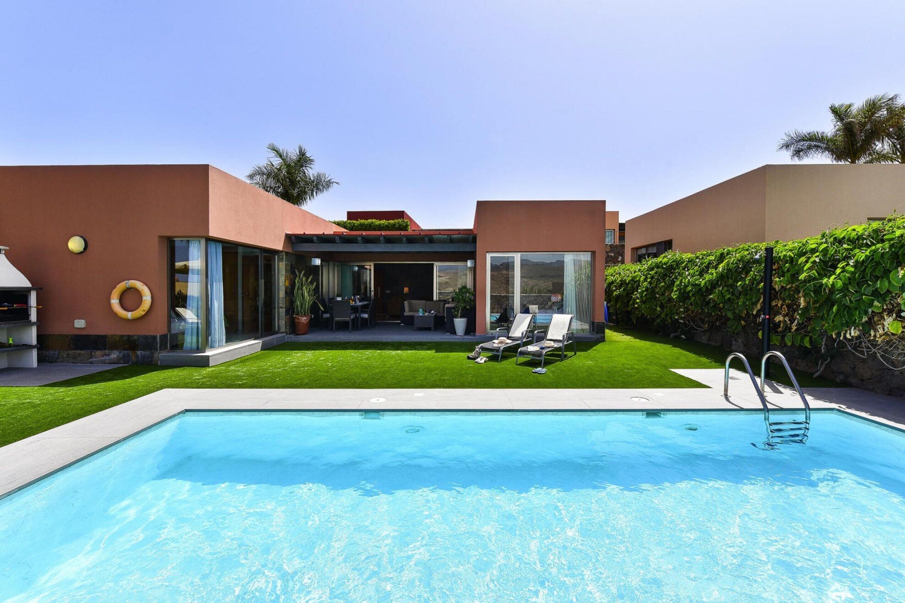 Uma área de piscina moderna com piscina azul cristalina, duas espreguiçadeiras e um pequeno espaço com cozinha ao ar livre.