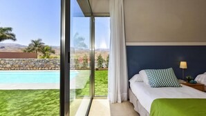 Une chambre avec une grande fenêtre, un lit recouvert d’une couverture verte, un oreiller à motifs et une table de chevet avec une lampe et une petite plante.