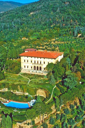A large, multi-story building with a red roof, surrounded by lush greenery and a swimming pool.