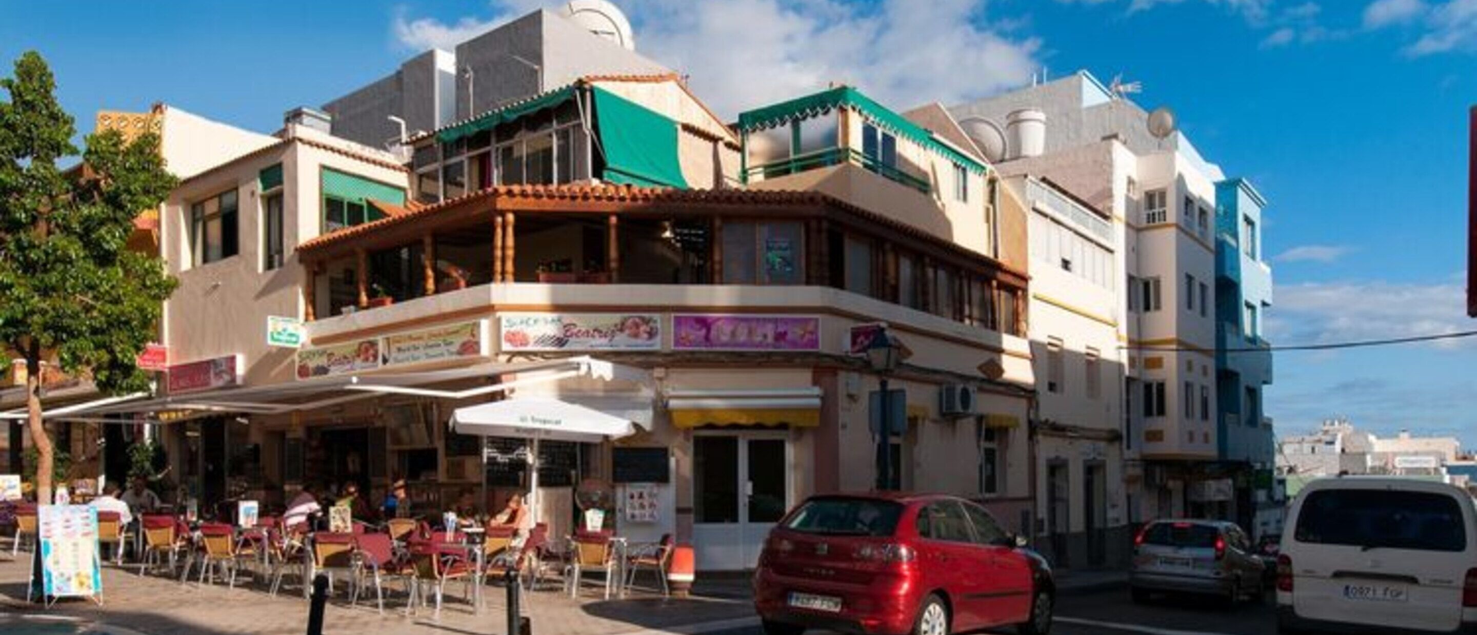 Une vue de rue avec un bâtiment doté d’une terrasse, une voiture rouge et un fourgon blanc.